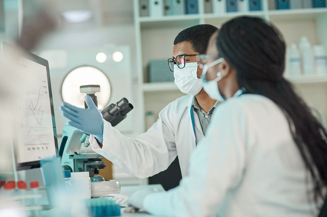 Two masked scientists in lab coats review data on a computer, microscope nearby